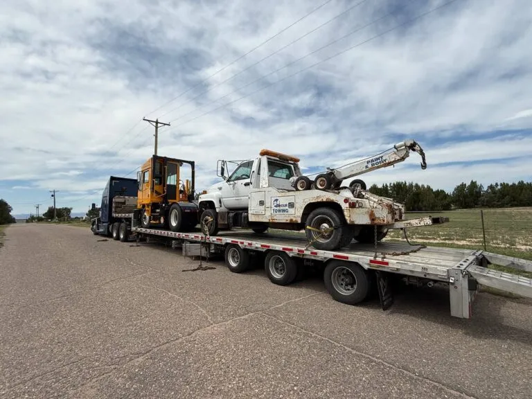 White tow truck and orange yard truck strapped down on a flatbed trailer hauled by a semi truck