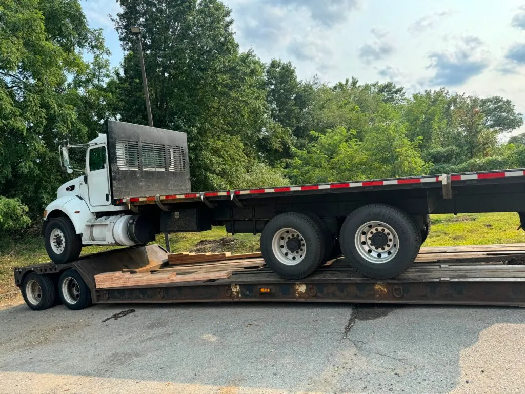 White flatbed truck loaded onto a lowboy trailer with wood blocking for heavy vehicle transport