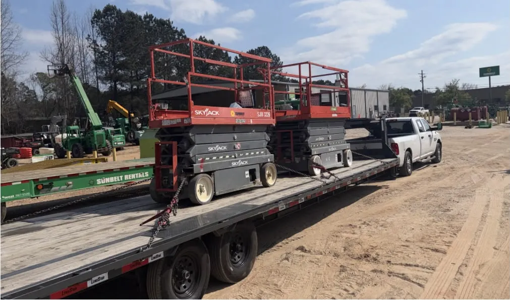 Two Skyjack scissor lifts chained to a gooseneck flatbed trailer behind a white pickup truck